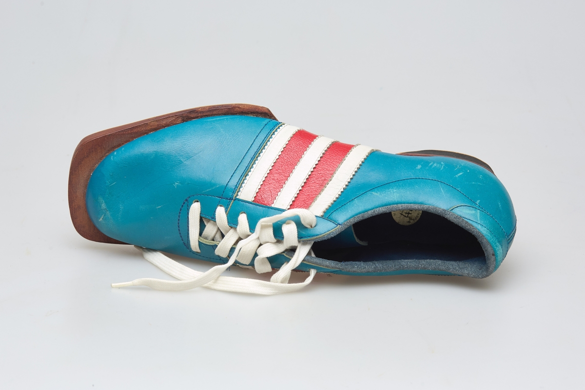 En skisko av turkisblått lær med røde og hvite striper på siden. Indre tunge og snøring med hvite skolisser. Beige såle av gummi.