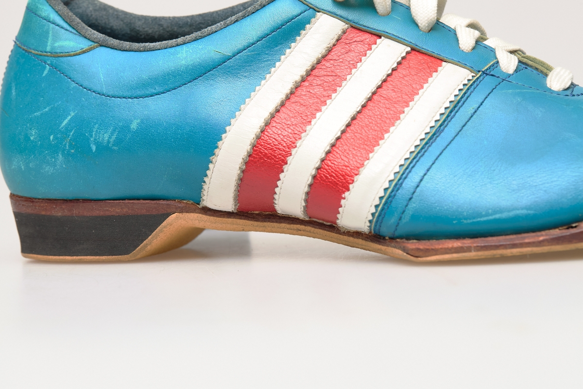 En skisko av turkisblått lær med røde og hvite striper på siden. Indre tunge og snøring med hvite skolisser. Beige såle av gummi.