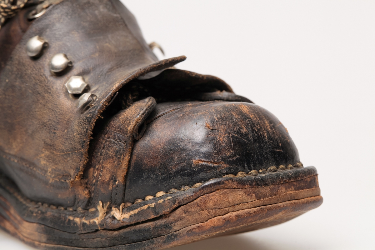 En skisko av brunt lær. Indre tunge og snøring gjennom maljer og hekter med skolisser. Overlappende lag med snøring utenpå dette, med hekter. Såle av lær.