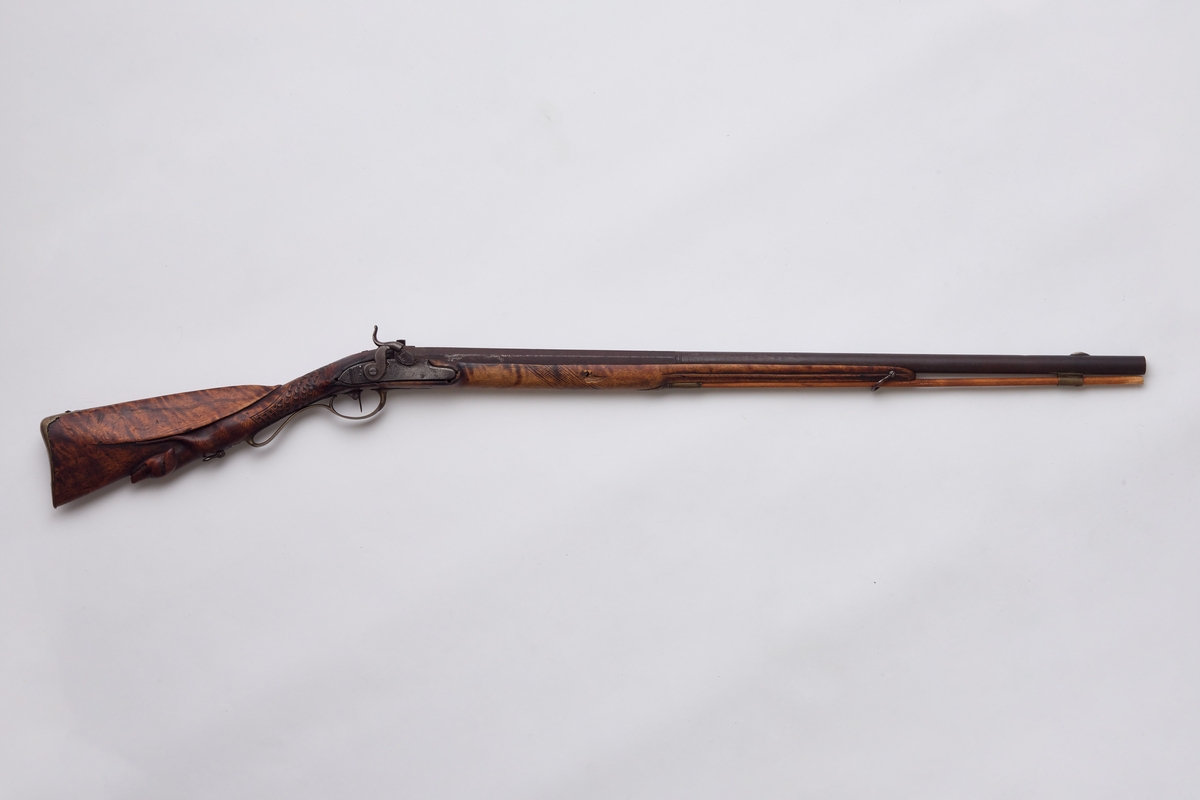 Gevær, glattløpet. Geværet har perkusjonslås, åttekantet pipe til 30,7 cm fra kammerenden, resten rund. Rikt utskåret skjefte, på låsblikket; S. Richards, og blomsterdekor bakerst. Geværet har kappe av messing med hæl og tå. Kolbehalsen er rikt skåret med striper og buer, og går rundt kolben over i en hund med framstrakt høyre ben og hengende ører. Fra spissrørkenet har forskjeftet profiler langsmed. Midtrørken mangler, det forreste er loddet til pipen. Under på pipens kammerende er innslått V. Påloddet sikteskur bakerst på pipen. Halvmåneformet 22 m. lang messingkorn med forreste ender 60 mm fra munningen. Låset har rik bladdekor på studdelen. Hanekroppen er S-formet og flat, hanemulen rund, hanefløyen glatt, med en liten bakoverbøy øverst. Lett jaktgevær.