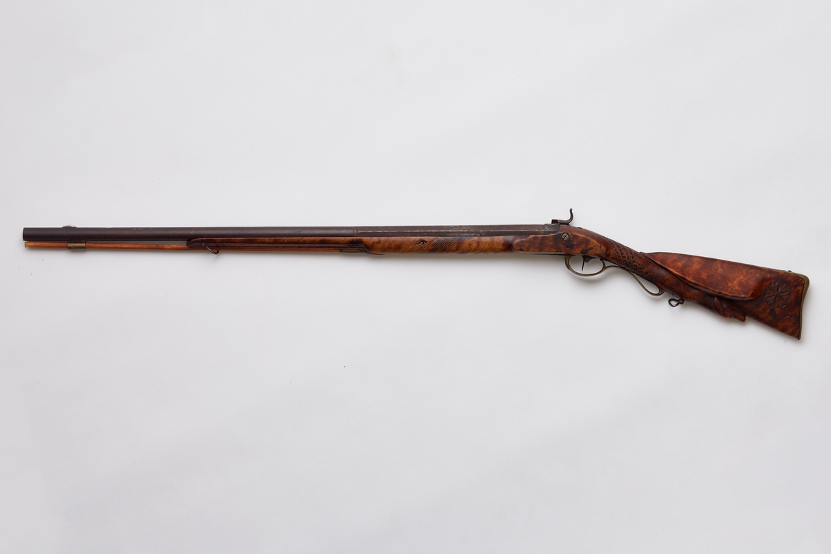 Gevær, glattløpet. Geværet har perkusjonslås, åttekantet pipe til 30,7 cm fra kammerenden, resten rund. Rikt utskåret skjefte, på låsblikket; S. Richards, og blomsterdekor bakerst. Geværet har kappe av messing med hæl og tå. Kolbehalsen er rikt skåret med striper og buer, og går rundt kolben over i en hund med framstrakt høyre ben og hengende ører. Fra spissrørkenet har forskjeftet profiler langsmed. Midtrørken mangler, det forreste er loddet til pipen. Under på pipens kammerende er innslått V. Påloddet sikteskur bakerst på pipen. Halvmåneformet 22 m. lang messingkorn med forreste ender 60 mm fra munningen. Låset har rik bladdekor på studdelen. Hanekroppen er S-formet og flat, hanemulen rund, hanefløyen glatt, med en liten bakoverbøy øverst. Lett jaktgevær.