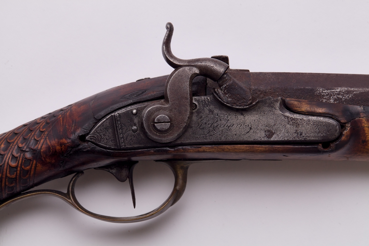 Gevær, glattløpet. Geværet har perkusjonslås, åttekantet pipe til 30,7 cm fra kammerenden, resten rund. Rikt utskåret skjefte, på låsblikket; S. Richards, og blomsterdekor bakerst. Geværet har kappe av messing med hæl og tå. Kolbehalsen er rikt skåret med striper og buer, og går rundt kolben over i en hund med framstrakt høyre ben og hengende ører. Fra spissrørkenet har forskjeftet profiler langsmed. Midtrørken mangler, det forreste er loddet til pipen. Under på pipens kammerende er innslått V. Påloddet sikteskur bakerst på pipen. Halvmåneformet 22 m. lang messingkorn med forreste ender 60 mm fra munningen. Låset har rik bladdekor på studdelen. Hanekroppen er S-formet og flat, hanemulen rund, hanefløyen glatt, med en liten bakoverbøy øverst. Lett jaktgevær.