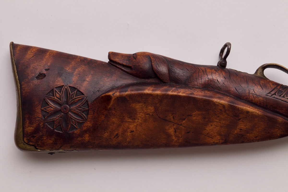 Gevær, glattløpet. Geværet har perkusjonslås, åttekantet pipe til 30,7 cm fra kammerenden, resten rund. Rikt utskåret skjefte, på låsblikket; S. Richards, og blomsterdekor bakerst. Geværet har kappe av messing med hæl og tå. Kolbehalsen er rikt skåret med striper og buer, og går rundt kolben over i en hund med framstrakt høyre ben og hengende ører. Fra spissrørkenet har forskjeftet profiler langsmed. Midtrørken mangler, det forreste er loddet til pipen. Under på pipens kammerende er innslått V. Påloddet sikteskur bakerst på pipen. Halvmåneformet 22 m. lang messingkorn med forreste ender 60 mm fra munningen. Låset har rik bladdekor på studdelen. Hanekroppen er S-formet og flat, hanemulen rund, hanefløyen glatt, med en liten bakoverbøy øverst. Lett jaktgevær.