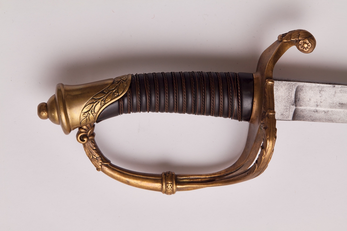 Sabel a): Klingen hulslipt til 17,5 cm fra spissen. Ryggen er tynnslipt fra spissen og 17 cm. oppover. Grepets plate, bøyle og knapp er støpt av messing. Kavlen er av presset horn med rifler hvori metallbånd. Dekorert.