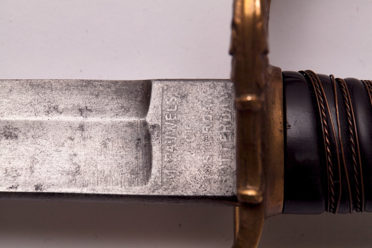 Sabel a): Klingen hulslipt til 17,5 cm fra spissen. Ryggen er tynnslipt fra spissen og 17 cm. oppover. Grepets plate, bøyle og knapp er støpt av messing. Kavlen er av presset horn med rifler hvori metallbånd. Dekorert.