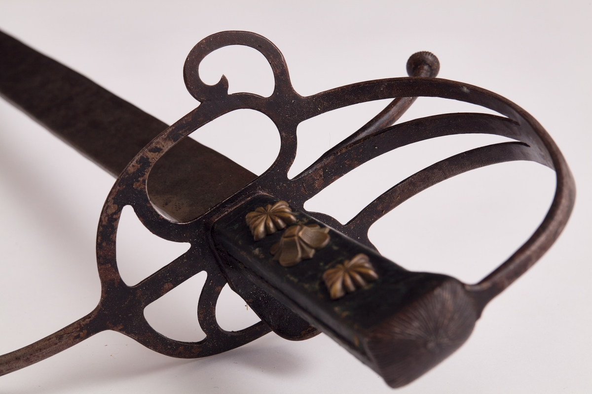 Sabel a): Klingen hulslipt til 17,5 cm fra spissen. Ryggen er tynnslipt fra spissen og 17 cm. oppover. Grepets plate, bøyle og knapp er støpt av messing. Kavlen er av presset horn med rifler hvori metallbånd. Dekorert.