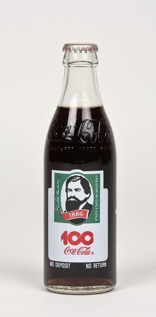 Läskedrycksflaska: Coca-Cola. Flaskan är tillverkad av klart glas och har två färgtryckta etiketter med text, varav en etikett har ett porträtt av John Pemberton - Coca-Colas grundare. I glaset finns två graveringar i form av Coca-Cola logotyper samt graverad text: "REGISTERED TRADE MARK". 296ml. Oöppnad.