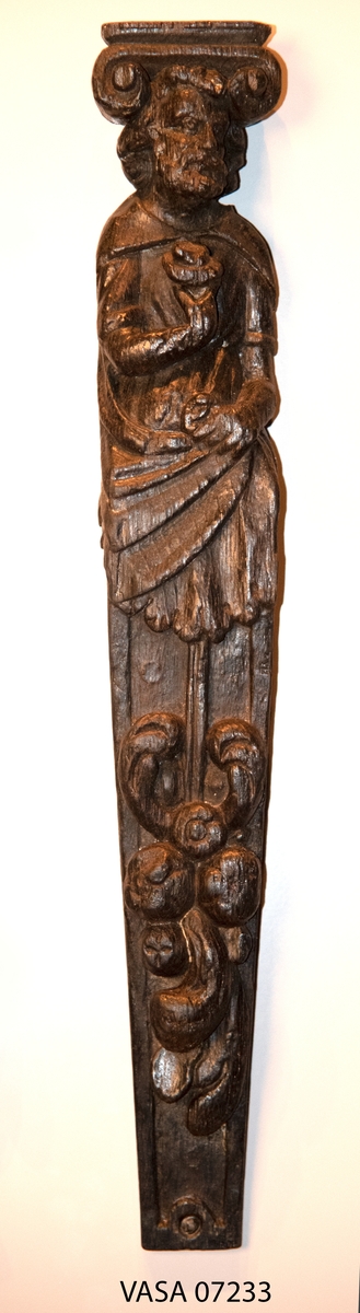 Hermpilaster.
Liten skulpterad pilaster föreställande skäggig herm under joniskt kapitäl.
Huvudet är vridet mot vänster. Hermen är insvept i en mantel som hålls fast med vänster hand framför magen. I höger hand håller figuren ett okänt föremål (blomsterkälk?) framför bröstet. Pilasterskaftet är prytt med stora frukter och blad, fasthängande vid en stängel som delvis täcker det nedåt avsmalnande skaftets ramverk. Baksidan är slät.
Skulpturen är mycket välbevarad med sju spikhål.

Text in English: Small sculpted pilaster with a bearded Herm beneath an Ionic capital.
The head of the figure is turned to the left. The Herm is enveloped in a mantle, held fast with the left hand in front of its stomach. Its right hand holds an unknown object (a bunch of flowers?) in front of its breast. The shaft of the pilaster is decked with large fruit and leaves hanging from a stalk which partly cover the framing of the tapering shaft. Its back is flat.
The sculpture is very well preserved.