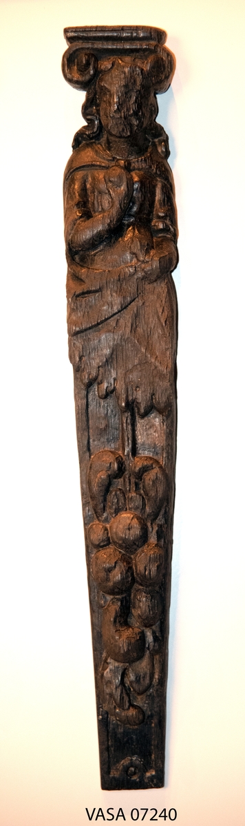 Hermpilaster.

Liten pilaster i form av en karyatid, med långt böljande hår under joniskt kapitäl.

Huvudet är vridet en aning åt höger. Med vänster hand hålls manteln samlad. I höger hand håller figuren en frukt eller dylikt framför bröstet. Pilasterskaftet är prytt med stora frukter och blad, fasthängande vid en stängel som delvis täcker det nedåt avsmalnande skaftets ramverk. Baksidan är slät.

Skulpturen är välbevarad, förutom ansiktet som är kraftigt slitet.

Fyra stycken spikhål, en övertäckt med en plugg.


Text in English: Sculpted small pilaster with a Caryatid with long wavy hair beneath an Ionic capital.

Its head is turned slightly to the right, its left hand holds its gathered mantle. Its right hand holds fruit or the like in front of its brests. The shaft of the pilaster is decorated with fruit and large leaves hanging from a stalk, which partly cover the framing of the tapering shaft. Its back is flat.

The sculpture is somewhat worn, its facial expression has been obliterated.