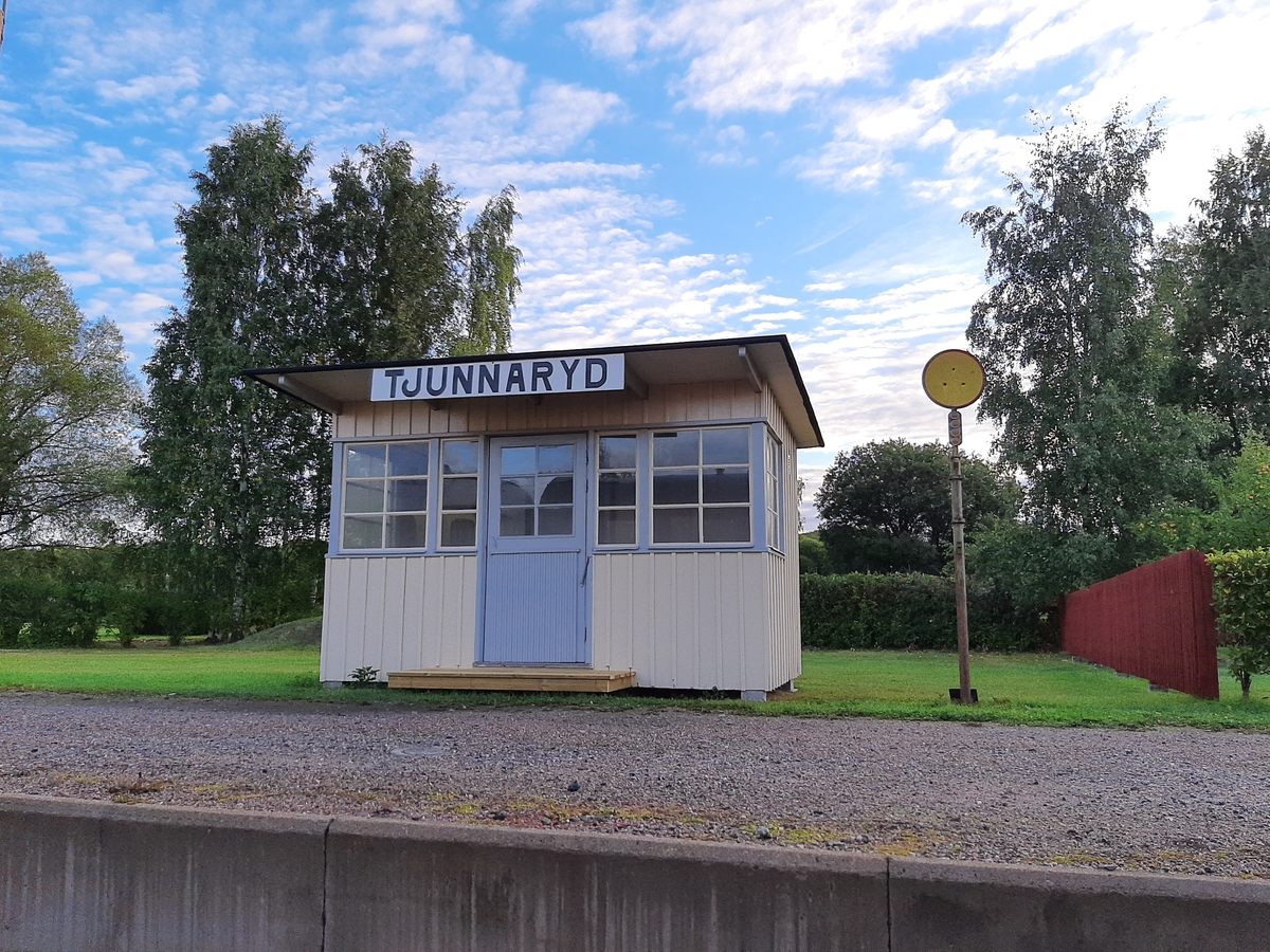 Hållplatskur från Tjunnaryd. Målad i ljusgul kulör med ljusgrå dörr, dörrfoder och fönsterfoder. Spröjsade fönster utmed hela framsidan, även på dörren, och delvis på sidorna.