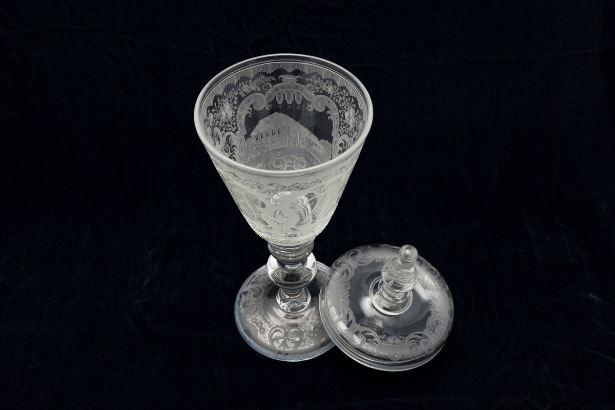 Pokal med lokk, glass. Fot har graverte blomster  og knekkede bånd. På cupa gravert bilde av Bogstad på en side, på den andre: N.W.J.  W.P.E.