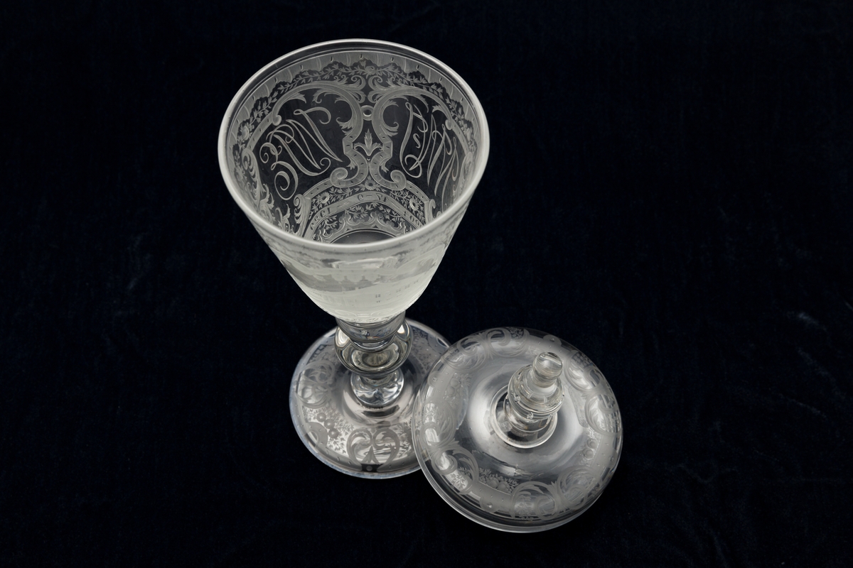 Pokal med lokk, glass. Fot har graverte blomster  og knekkede bånd. På cupa gravert bilde av Bogstad på en side, på den andre: N.W.J.  W.P.E.