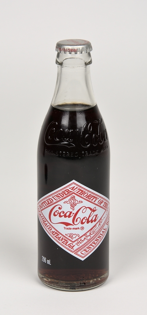 Läskedrycksflaska: Coca-Cola. Flaskan är tillverkad av klart glas och har två färgtryckta etiketter med text, varav en etikett har ett porträtt av John Pemberton - Coca-Colas grundare. I glaset finns två graveringar i form av Coca-Cola logotyper samt graverad text: "REGISTERED TRADE MARK". 296ml. Oöppnad.