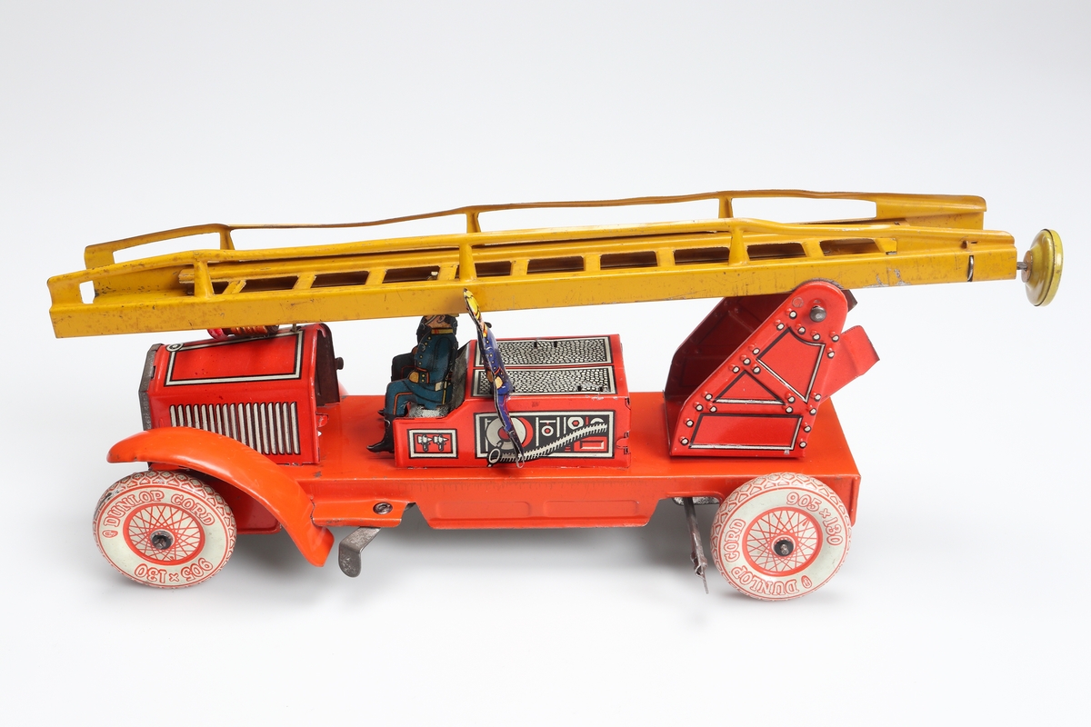 Leksaksbrandbil, tillverkad av tunn pressad plåt. Bilen är lackerad i rött med gulvita hjul, som har röda markeringar. Gul stege, som är hopskjutbar genom en lång, spiralvriden stång med en ratt i bakänden. Nitskallar, gälar på motorhuven och slangutrustning m.m. är markerade med svart och vit färg. Tre brandsoldater finns med (två saknas). De har blå uniformer, svarta stövlar och gula hjälmar. Två sitter på förarsätet och en på sidan, kylarmaskeringen är silverfärgad. Brandbilen drivs genom ett fjäderverk, vars nyckel sticker ut vid vänster bakhjul. Ovanpå motorhuven sitter en lamphållare med en rödmålad glödlampa i. Batteriet skall förvaras under bilen, i en särskild hållare, försedd med strömbrytare.