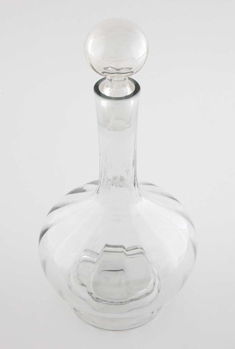 Glasskaraffel med propp i klart glass. Korpus er kuleformet med en lang og slank hals. Karaffelens riller snor seg fra korpus og oppover halsen.
