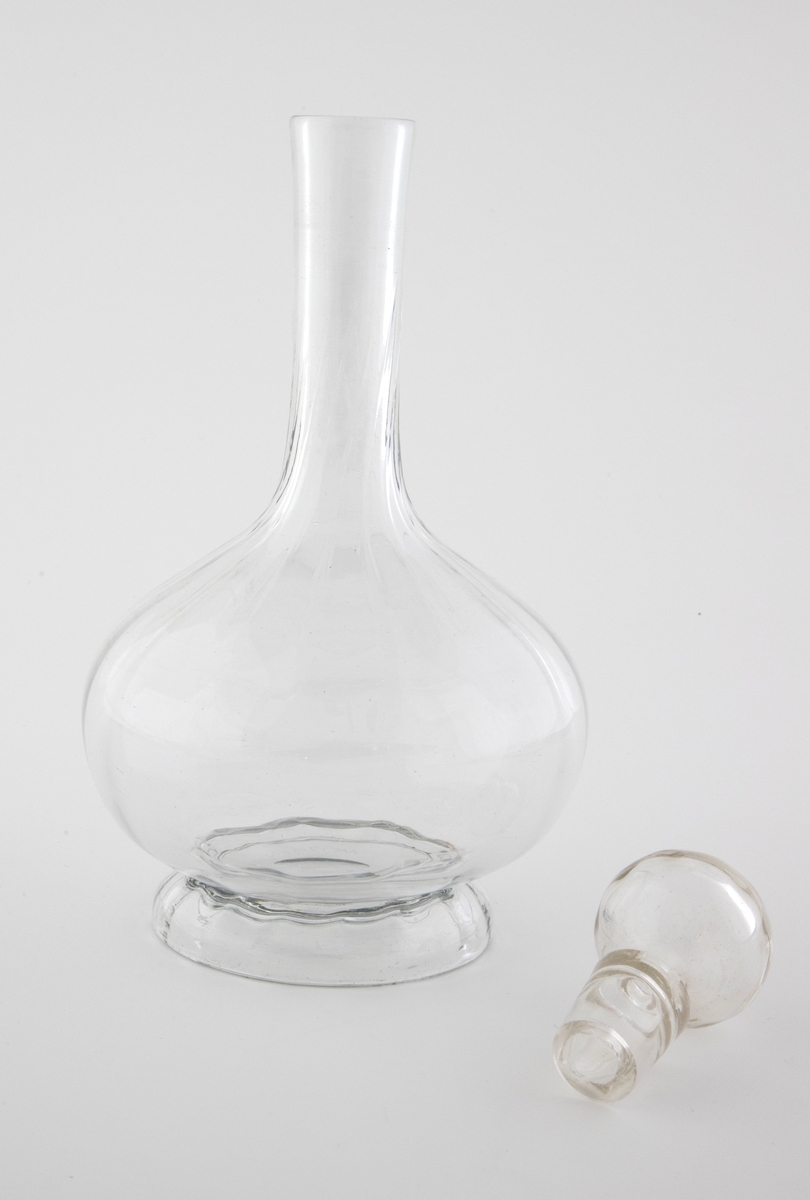 Glasskaraffel med propp i klart glass. Korpus er kuleformet med en lang og slank hals. Karaffelens riller snor seg fra korpus og oppover halsen.