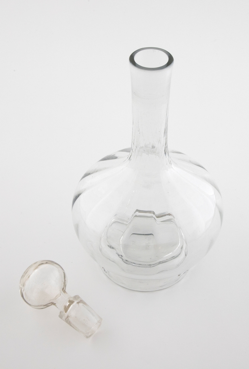 Glasskaraffel med propp i klart glass. Korpus er kuleformet med en lang og slank hals. Karaffelens riller snor seg fra korpus og oppover halsen.