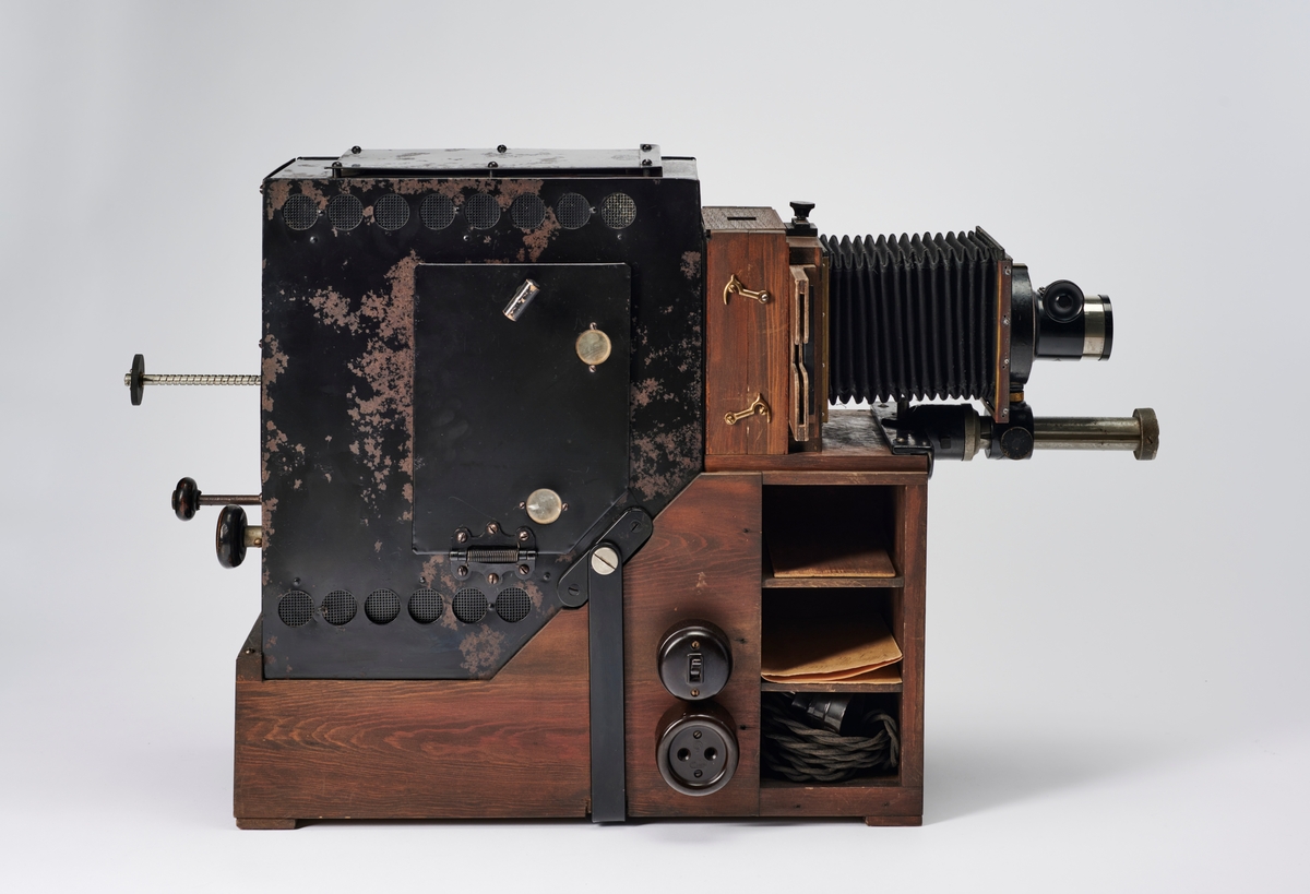 Lysbildefremviser med elektrisk lyskilde, bygd av arkitekt Hans Grendahl (1877-1957) ved hjelp av både kommersielle og egenkonstruerte deler. Grendahl har med dette projeksjonsapparatet forbedret en Skioptikon fra ukjent fabrikant og satt på en del med kondensatorlinse (to halvkonvekslinser) for å fordele lyset bedre med mulighet til å åpne for rengjøring, belg og en projeksjonslinse. På baksiden av apparatet stilles intensiteten til den elektriske lyspæren som befinner seg inne i lampehuset.
Objektivet er et Tessar 1:4,5, f= 18 cm DPR Carl Zeiss Jena - Nr. 220783 objektiv.