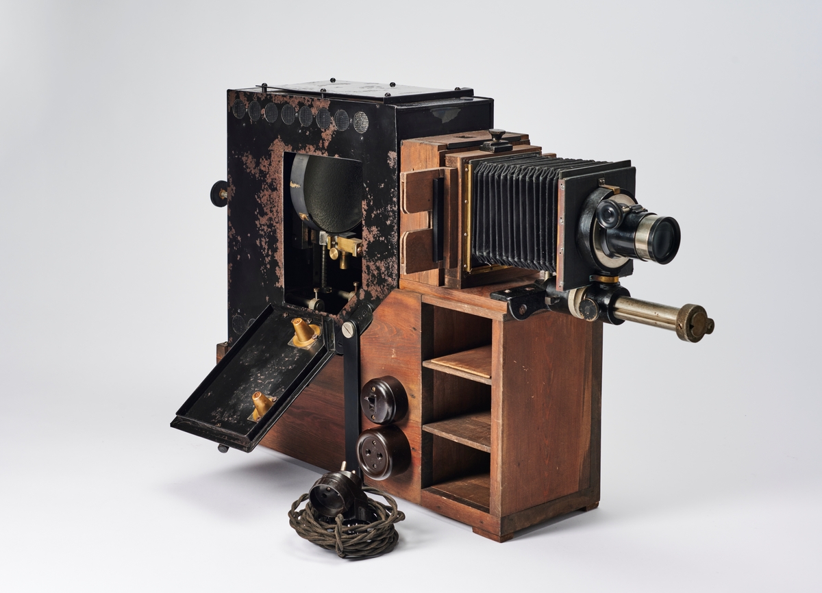 Lysbildefremviser med elektrisk lyskilde, bygd av arkitekt Hans Grendahl (1877-1957) ved hjelp av både kommersielle og egenkonstruerte deler. Grendahl har med dette projeksjonsapparatet forbedret en Skioptikon fra ukjent fabrikant og satt på en del med kondensatorlinse (to halvkonvekslinser) for å fordele lyset bedre med mulighet til å åpne for rengjøring, belg og en projeksjonslinse. På baksiden av apparatet stilles intensiteten til den elektriske lyspæren som befinner seg inne i lampehuset.
Objektivet er et Tessar 1:4,5, f= 18 cm DPR Carl Zeiss Jena - Nr. 220783 objektiv.