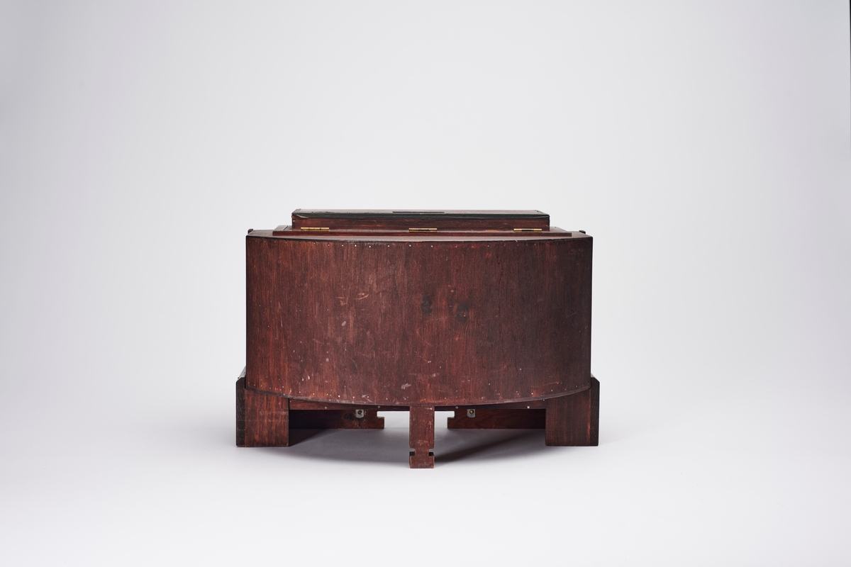 Lampehus til forstørrelsesapparat, konstruert av arkitekt H. Grendahl (1877-1957) i 1931. Det har plass til glassplater, to elektriske lyskilder og en reflektor, som gir et indirekte lys. En slik indirekte lyskilde sparer glassplatene for varme, da det ikke blir like varmt som ved direkte lys. Apparatet har flere lystette ventilasjonsluker.