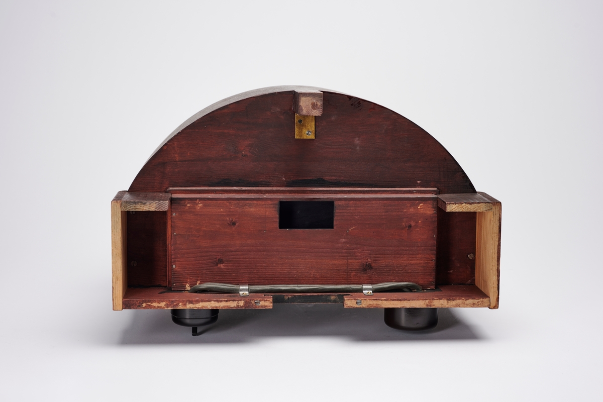 Lampehus til forstørrelsesapparat, konstruert av arkitekt H. Grendahl (1877-1957) i 1931. Det har plass til glassplater, to elektriske lyskilder og en reflektor, som gir et indirekte lys. En slik indirekte lyskilde sparer glassplatene for varme, da det ikke blir like varmt som ved direkte lys. Apparatet har flere lystette ventilasjonsluker.