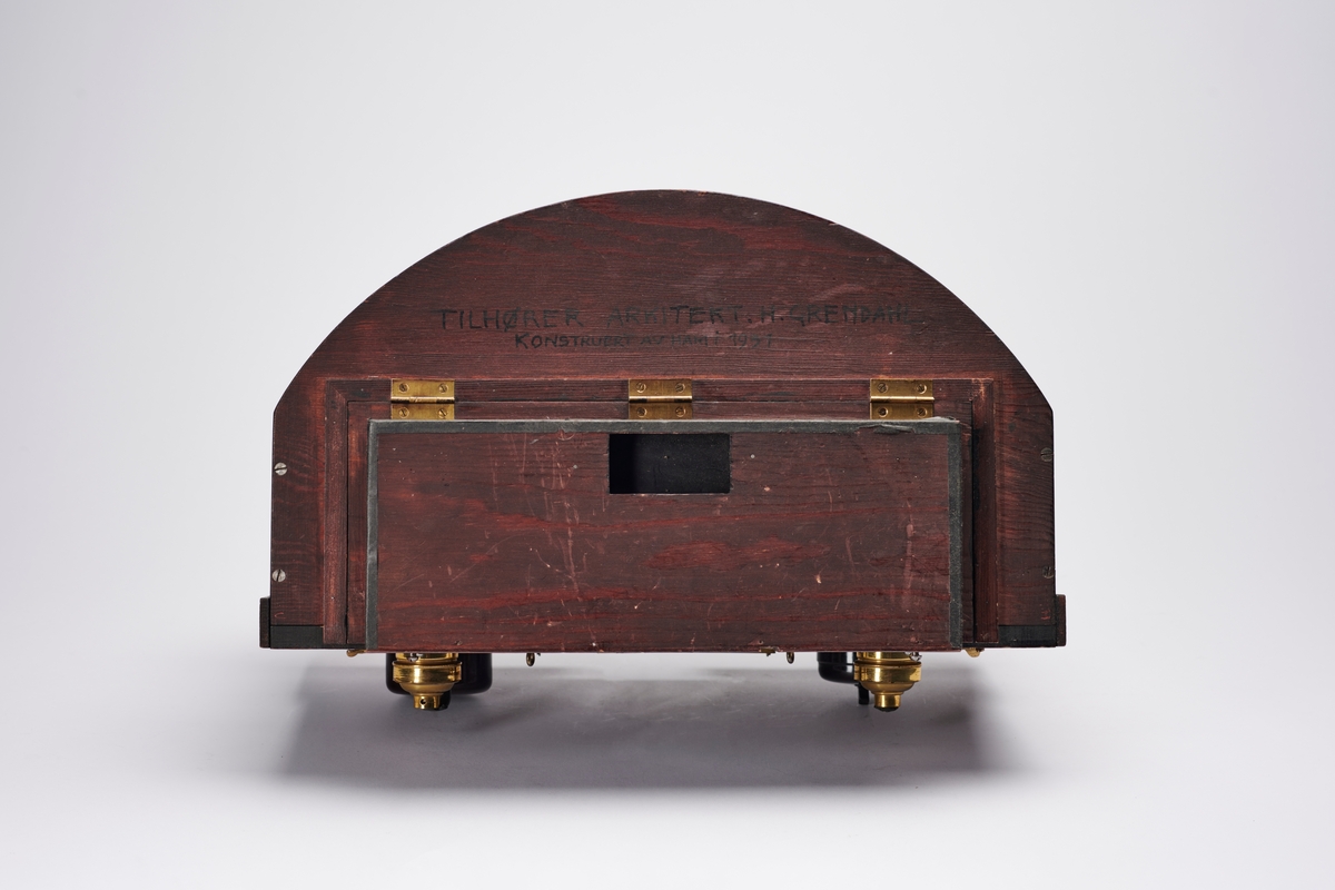Lampehus til forstørrelsesapparat, konstruert av arkitekt H. Grendahl (1877-1957) i 1931. Det har plass til glassplater, to elektriske lyskilder og en reflektor, som gir et indirekte lys. En slik indirekte lyskilde sparer glassplatene for varme, da det ikke blir like varmt som ved direkte lys. Apparatet har flere lystette ventilasjonsluker.
