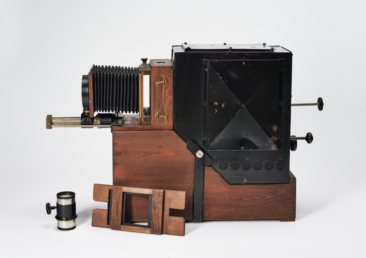 Lysbildefremviser med elektrisk lyskilde, bygd av arkitekt Hans Grendahl (1877-1957) ved hjelp av både kommersielle og egenkonstruerte deler. Grendahl har med dette projeksjonsapparatet forbedret en Skioptikon fra ukjent fabrikant og satt på en del med kondensatorlinse (to halvkonvekslinser) for å fordele lyset bedre med mulighet til å åpne for rengjøring, belg og en projeksjonslinse. På baksiden av apparatet stilles intensiteten til den elektriske lyspæren som befinner seg inne i lampehuset.
Objektivet er et Tessar 1:4,5, f= 18 cm DPR Carl Zeiss Jena - Nr. 220783 objektiv.