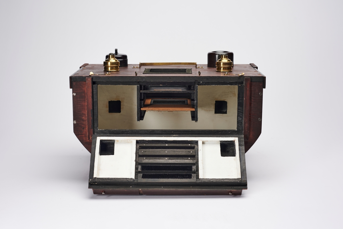 Lampehus til forstørrelsesapparat, konstruert av arkitekt H. Grendahl (1877-1957) i 1931. Det har plass til glassplater, to elektriske lyskilder og en reflektor, som gir et indirekte lys. En slik indirekte lyskilde sparer glassplatene for varme, da det ikke blir like varmt som ved direkte lys. Apparatet har flere lystette ventilasjonsluker.