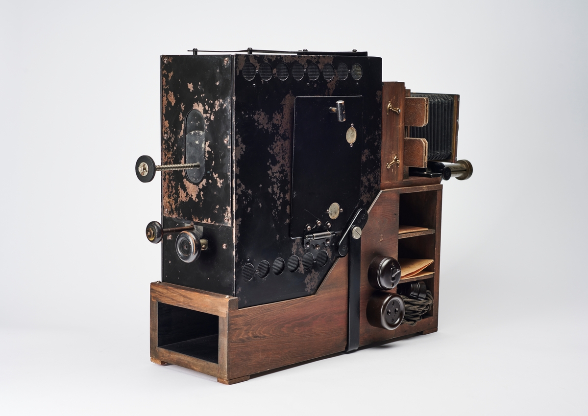 Lysbildefremviser med elektrisk lyskilde, bygd av arkitekt Hans Grendahl (1877-1957) ved hjelp av både kommersielle og egenkonstruerte deler. Grendahl har med dette projeksjonsapparatet forbedret en Skioptikon fra ukjent fabrikant og satt på en del med kondensatorlinse (to halvkonvekslinser) for å fordele lyset bedre med mulighet til å åpne for rengjøring, belg og en projeksjonslinse. På baksiden av apparatet stilles intensiteten til den elektriske lyspæren som befinner seg inne i lampehuset.
Objektivet er et Tessar 1:4,5, f= 18 cm DPR Carl Zeiss Jena - Nr. 220783 objektiv.