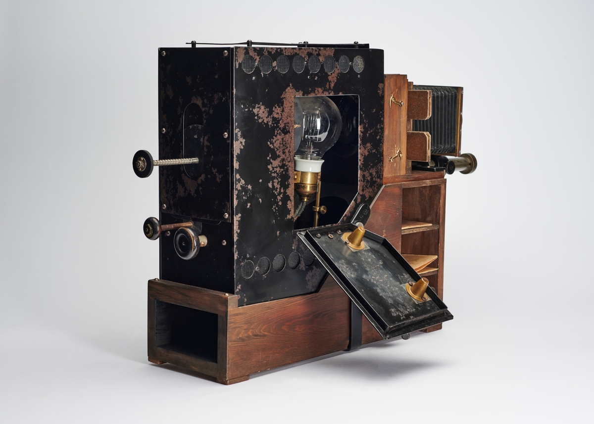 Lysbildefremviser med elektrisk lyskilde, bygd av arkitekt Hans Grendahl (1877-1957) ved hjelp av både kommersielle og egenkonstruerte deler. Grendahl har med dette projeksjonsapparatet forbedret en Skioptikon fra ukjent fabrikant og satt på en del med kondensatorlinse (to halvkonvekslinser) for å fordele lyset bedre med mulighet til å åpne for rengjøring, belg og en projeksjonslinse. På baksiden av apparatet stilles intensiteten til den elektriske lyspæren som befinner seg inne i lampehuset.
Objektivet er et Tessar 1:4,5, f= 18 cm DPR Carl Zeiss Jena - Nr. 220783 objektiv.