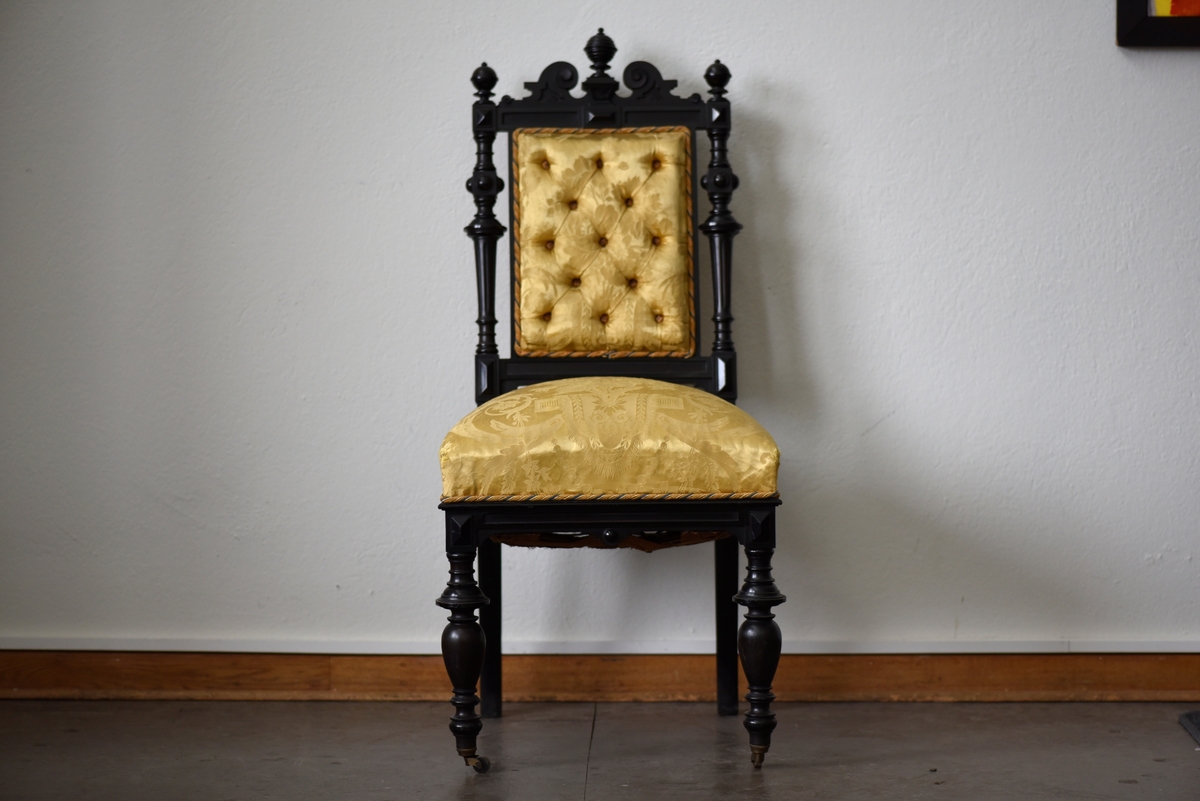 Stol i nyrenässansstil tillverkad av svart päronträ och klädd i gul sidendamast. 
Sargen och ryggstödsramen är rikt utsnidade. Det främre benparet är svarvade och försedda med trissor. Det bakre benparet är skurna, något svängda och bakåtlutande. 
Den stoppade sitsen och ryggbrickan är klädda med ett gult sidendamasttyg med invävt mönster i form av blommor, blad och band bland annat. Ryggbrickan är försedd med 13 klädda knappar.
Runt klädselns kanter mot ramverket sitter ett tvinnat kantband i gult, vitt och blått.