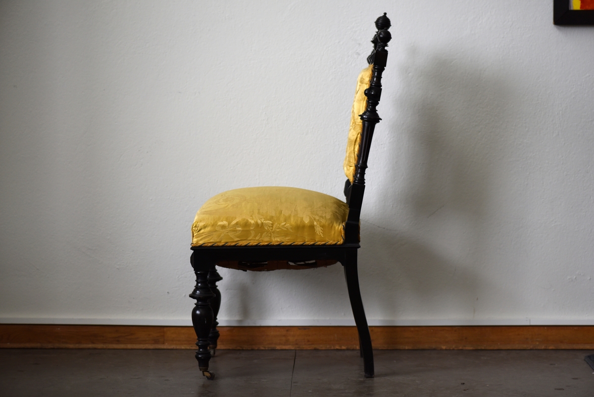 Stol i nyrenässansstil tillverkad av svart päronträ och klädd i gul sidendamast. 
Sargen och ryggstödsramen är rikt utsnidade. Det främre benparet är svarvade och försedda med trissor. Det bakre benparet är skurna, något svängda och bakåtlutande. 
Den stoppade sitsen och ryggbrickan är klädda med ett gult sidendamasttyg med invävt mönster i form av blommor, blad och band bland annat. Ryggbrickan är försedd med 13 klädda knappar.
Runt klädselns kanter mot ramverket sitter ett tvinnat kantband i gult, vitt och blått.
