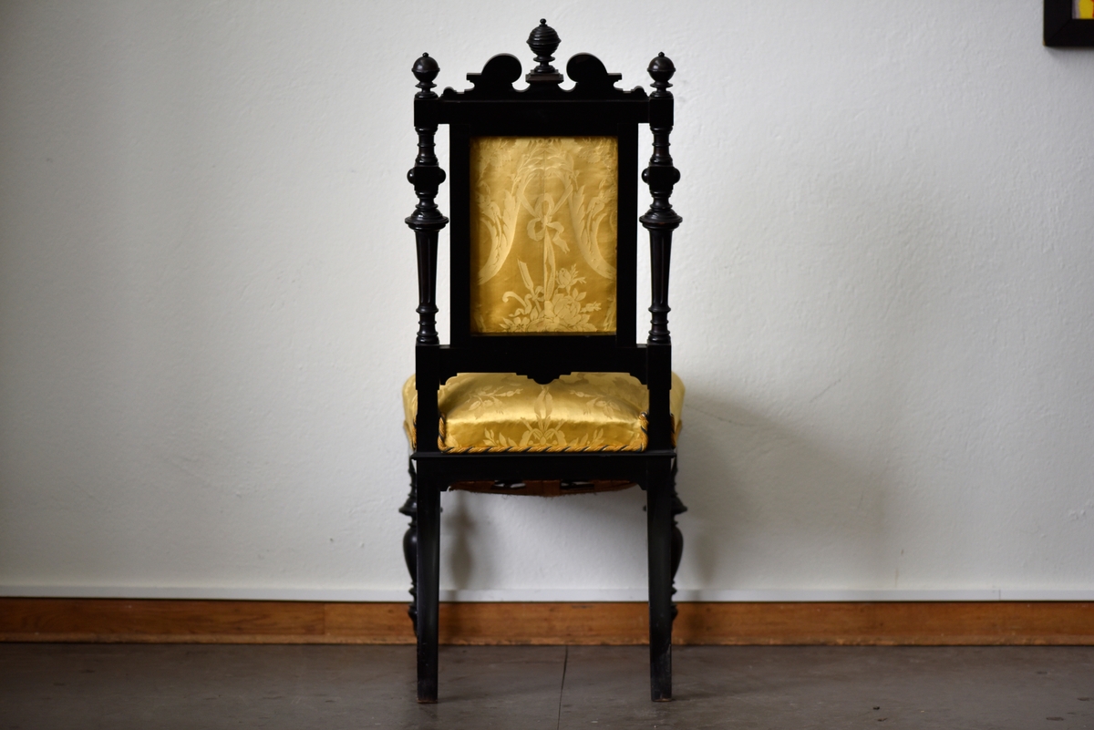 Stol i nyrenässansstil tillverkad av svart päronträ och klädd i gul sidendamast. 
Sargen och ryggstödsramen är rikt utsnidade. Det främre benparet är svarvade och försedda med trissor. Det bakre benparet är skurna, något svängda och bakåtlutande. 
Den stoppade sitsen och ryggbrickan är klädda med ett gult sidendamasttyg med invävt mönster i form av blommor, blad och band bland annat. Ryggbrickan är försedd med 13 klädda knappar.
Runt klädselns kanter mot ramverket sitter ett tvinnat kantband i gult, vitt och blått.
