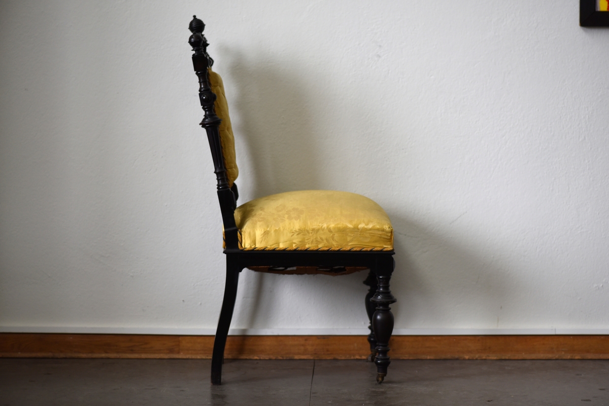 Stol i nyrenässansstil tillverkad av svart päronträ och klädd i gul sidendamast. 
Sargen och ryggstödsramen är rikt utsnidade. Det främre benparet är svarvade och försedda med trissor. Det bakre benparet är skurna, något svängda och bakåtlutande. 
Den stoppade sitsen och ryggbrickan är klädda med ett gult sidendamasttyg med invävt mönster i form av blommor, blad och band bland annat. Ryggbrickan är försedd med 13 klädda knappar.
Runt klädselns kanter mot ramverket sitter ett tvinnat kantband i gult, vitt och blått.
