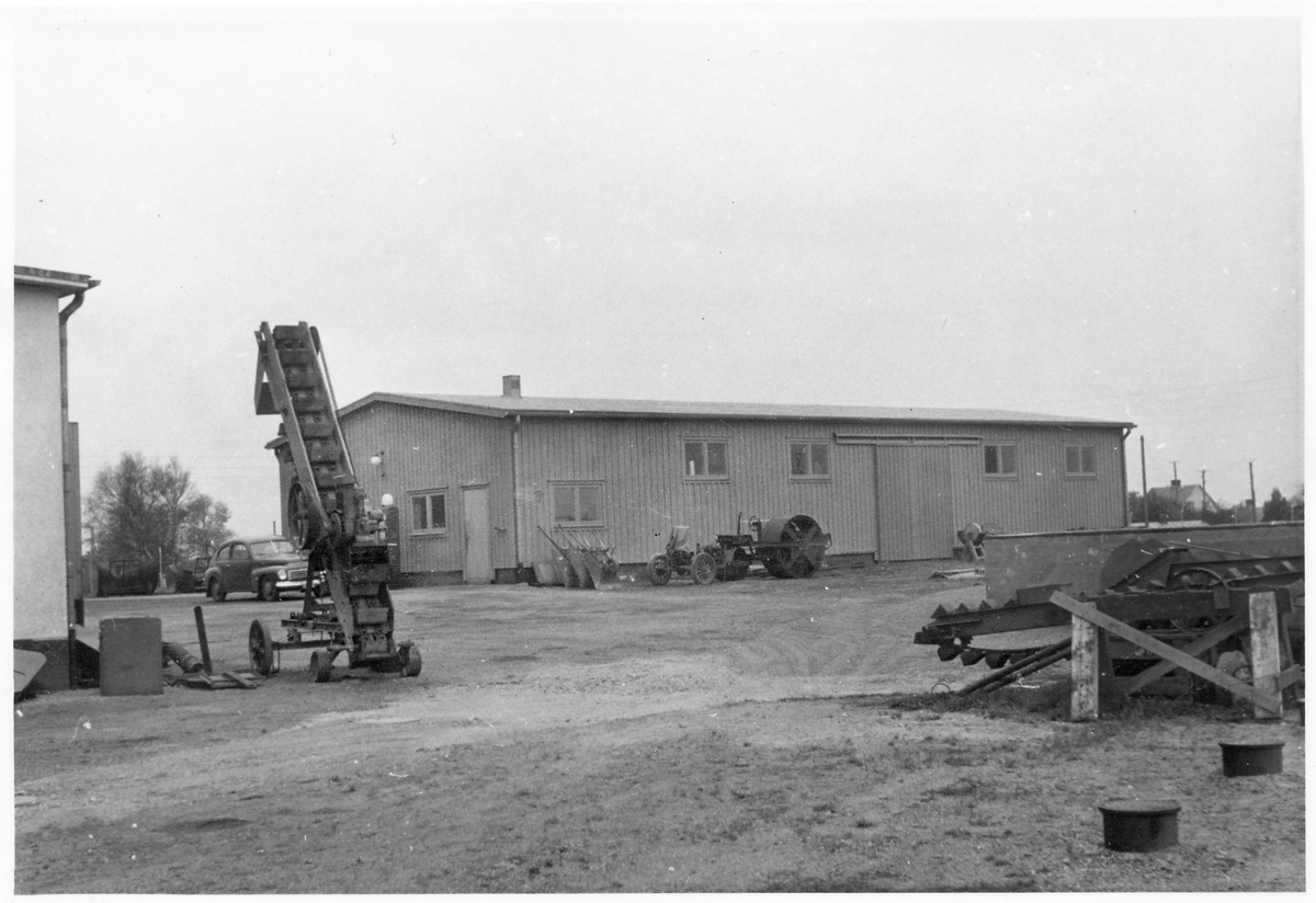 Vägstation N6, Varberg. Garageplan med förrådsbyggnad. Framför byggnaden asfaltvält Borås mekaniska verkstad, till vänster skopelevator, lastningselevator. Skottkärror lutade mot väggen. Slipstenbock med slipsten. Vid gaveln bensinpumpar (drivmedelspumpar) och personbil Volvo PV.