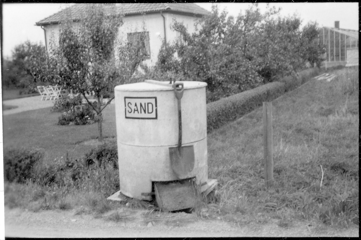 Vägstation P12, Åsaka. Sandlåda, eventuellt av cement. Spade, öppning nertill för hämtning av sanden. Skylt "Sand".