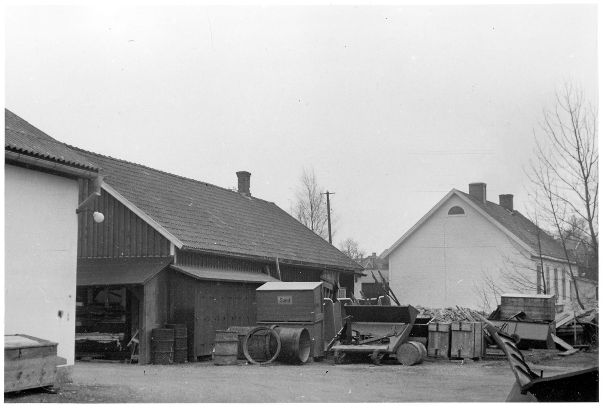 Vägstation P15, Tösse, filial Mellerud. Förrådsbyggnader med diverse föremål och material framför, bland annat sandspridare, typ "Sprider" tillverkad av Trafik & Vägmärken, Stockholm, sandlådor med skylten "Sand", verktygslådor. Mot väggen manskapsskåp för manskapets personliga utrustning och redskap. 