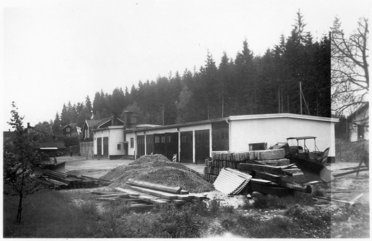 Vägstation P16, Bengtsfors. Vägstationens huvudbyggnad med verkstad, kontor och garage. Bortom denna byggnad äldre byggnad med skylt på framsidan "Vedbo vägdistrikt". Upplag av grus, kantsten, järnbalkar med mera på garageplan. Väghyvel och spetsplog.
