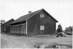 Vägstation R10, Kålltorp. Garagebyggnad och förrådsbyggnad. Bakom byggnaden personbil Volvo PV och traktor, upplag av cementrör. Mellan byggnaderna en bensinpump (drivmedelspump). Invid väg.