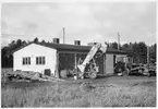 Vägstation R11, Grästorp. Garagebyggnad. Framför byggnaden lastningselevator (skopelevator), väghyvel, eventuell Svedala typ BD2. Smörjbrygga vid gaveln. Lösa hjul till väghållningsmaskiner.