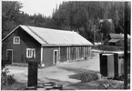 Vägstation S5, Sysslebäck. Garagebyggnad. I förgrunden bensinpump (drivmedelspump), till höger manskapsvagn, ÅSA transportabla manskapsbod, Åmåls Sågverks AB (1940-talet). Bostadshus i bakgrunden.