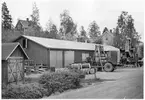 Vägstation S12, Åmotfors. Förrådsbyggnad. Framför byggnaden väghyvel, bortom denna en lastningselevator (skopelevator). Upplag av bränslefat. Bostadshus i bakgrunden. Invid väg.