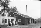 Vägstation W9, Nås, filial Dala-Järna. Garagebyggnad. Cyklar och motorcykel vid garagebyggnadens gavel. Bensinpump (drivmedelspump) i bakgrunden. Garagebyggnaden invid bensinstation (bensinmack) med tre pumpar. Bensinstationens servicebyggnad hopbyggd med bostadshus och garagebyggnaden. På taket av bensinstationens servicebyggnad skylt 