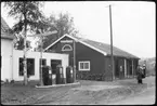 Vägstation W9, Nås, filial Dala-Järna.  Garagebyggnad. Cyklar och motorcykel vid garagebyggnadens gavel. Bensinpump (drivmedelspump) i bakgrunden. Garagebyggnaden invid bensinstation (bensinmack) med tre pumpar. Bensinstationens servicebyggnad hopbyggd med bostadshus och garagebyggnaden. På taket av bensinstationens servicebyggnad skylt 