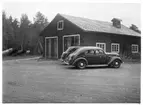 Vägstation W13, Älvdalen. Garagebyggnad med portar på gaveln. Bakom byggnaden sten- och stubbrytare, spetsplogar. Två personbilar på garageplan, en med VoV-emblem på dörren.