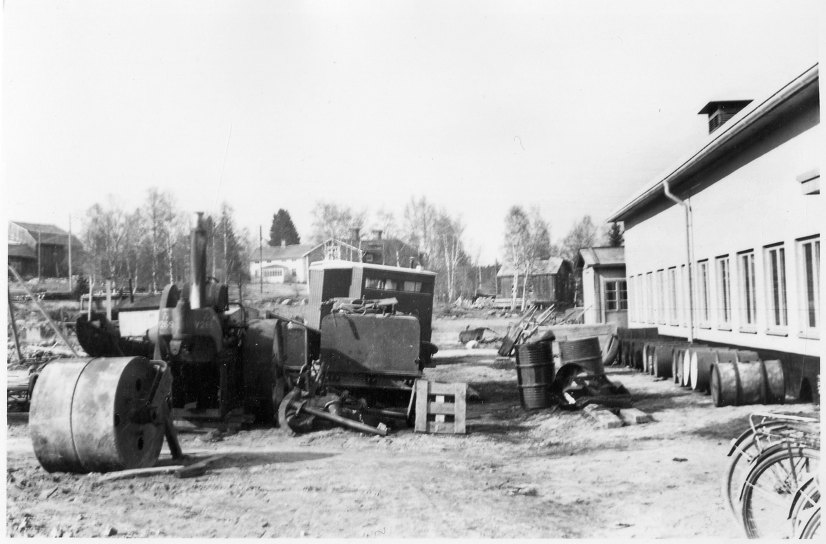 Vägstation Y6, Älandsbro. Baksidan av garagebyggnaden. Diverse material, "skrot". Motorvält Munktell, med VoV-maskinnummer V269, demonterad, främre valsen saknas. Vid vägväsendets förstatligande 1944 tillhörde välten Medelpads mellersta vägdistrikt. Manskapsvagn, ÅSA transportabla manskapsbod, Åmåls Sågverks AB, 1940-talet. Bostadshus i bakgrunden. Eventuellt depåverkstadens (länsverkstadens) material. 