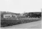 Vägstation Y11, Nyland. Vy över vägstationens huvudbyggnad, med från vänster verkstad, kontor och garage. Personbil, spetsplogar och sidoplogar på garageplan. Bensinpump (drivmedelspump). Framför verkstaden lastningselevator. Större magasinsbyggnad i bakgrunden. Invid väg.