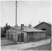 Y län. Före detta hamnförrådet. Förråds- och garagebyggnad, eventuellt också smedja,  vid infarten, med grind, till området. Cykel lutad mot telefonstolpe.