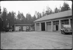 Vägstation AC4, Vännäs. Vägstationens huvudbyggnad, med garagedelen skymtar till höger. Förrådsbyggnad i bakgrunden. Personbil på garageplan, släpkärra till lastbil.