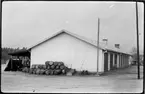 Vägstation AC 16, Åsele. Förråds- och garagebyggnad. Upplag av bränslefat vid gavel, skottkärra.